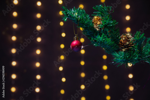 There is red ball on the green xmas tree branch. There are glowing lights/bokeh and on the background. Merry Christmas. Happy New Year 2020.  © Iakov