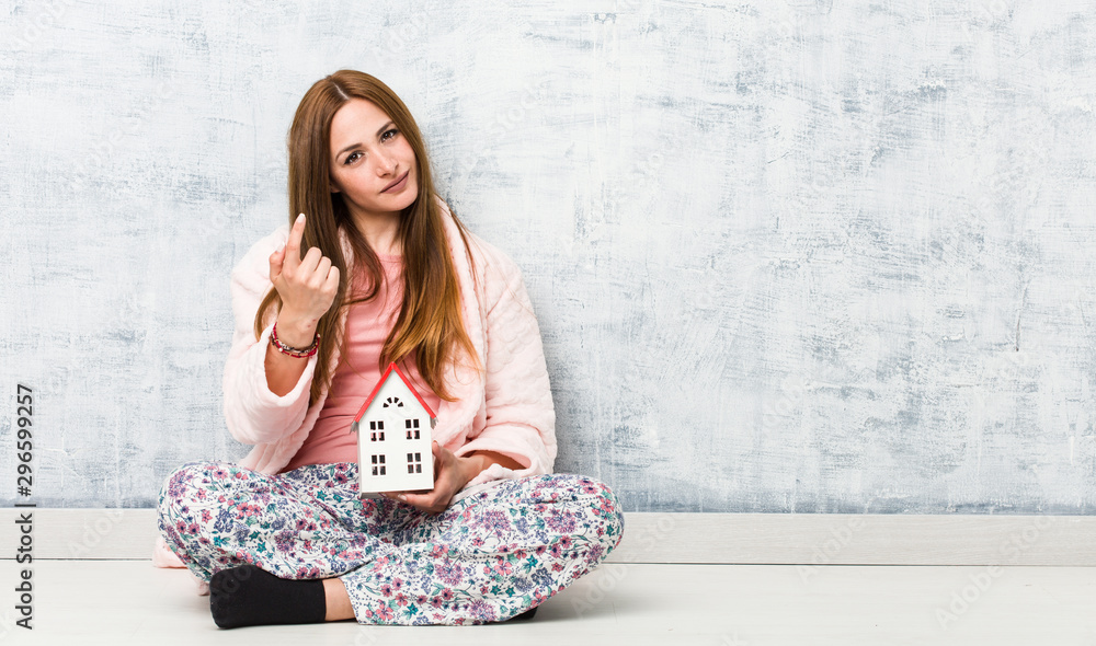 Young caucasian woman holding a house icon pointing with finger at you as if inviting come closer. Young caucasian woman holding a house icon pointing with finger at you as if inviting come closer.