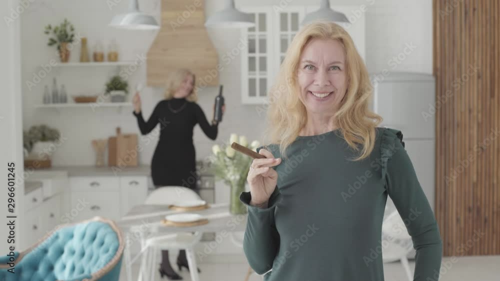 Rich mature Caucasian woman with a cigar scaterring money and smiling. Her positive friend dacing with wine and glasses at the background. Two wealthy senior ladies resting in the luxurious kitchen. Rich mature Caucasian woman with a cigar scaterring money and smiling. Her positive friend dacing with wine and glasses at the background. Two wealthy senior ladies resting in the luxurious kitchen.