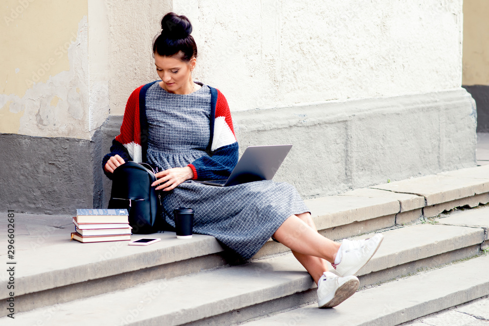 Female student is putting something to the backpack on the steps of university. Student girl looking for something in a backpack on the stairs. Female student is putting something to the backpack on the steps of university. Student girl looking for something in a backpack on the stairs.