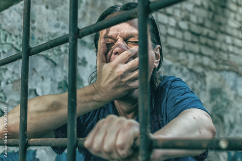 A depressed man holds his hands on the prison bars. Prisoner for the crime. Poor conditions. Scream, despair, pain, hunger.