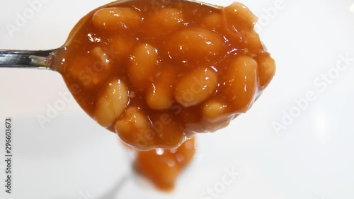 Video close up of metal spoon with British baked beans. Moving spoon and dropping baked beans on white plate. British comfort food. Haricot beans, also known as navy beans, in tomato sauce