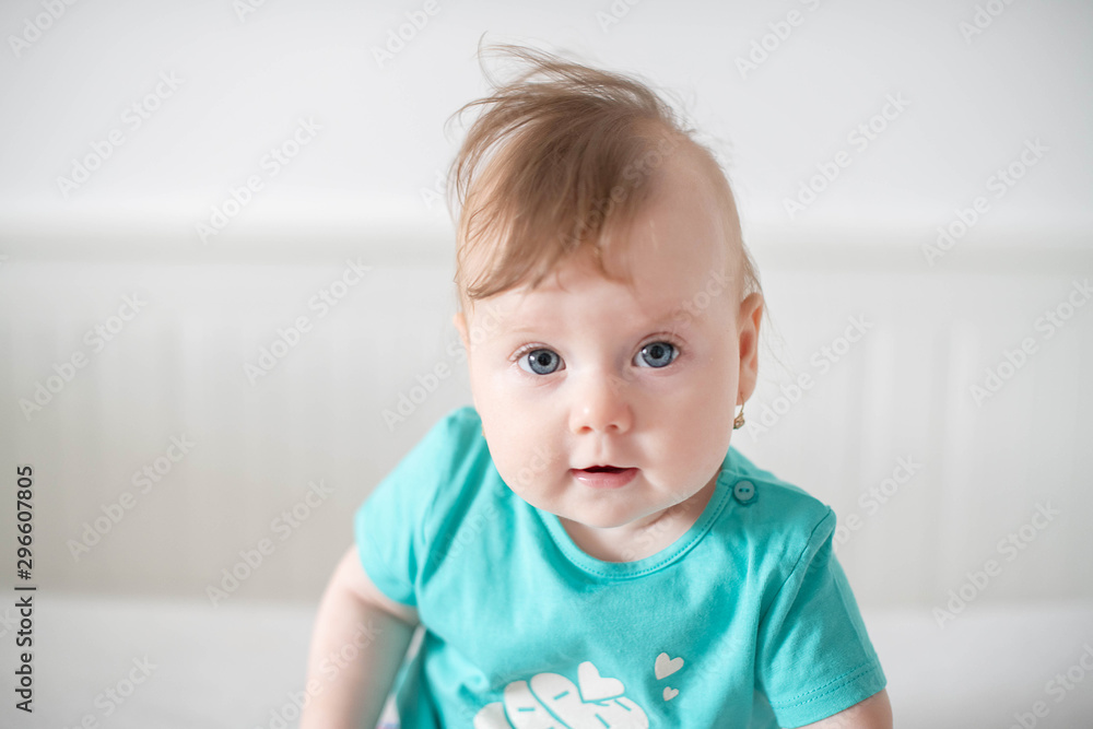 Portrait of adorable Caucasian baby girl with blue eyes, looking at the camera calmly, with curiosity, interactivity or inquisitiveness and sitting in a white baby cot; cute baby expressions concepts Portrait of adorable Caucasian baby girl with blue eyes, looking at the camera calmly, with curiosity, interactivity or inquisitiveness and sitting in a white baby cot; cute baby expressions concepts
