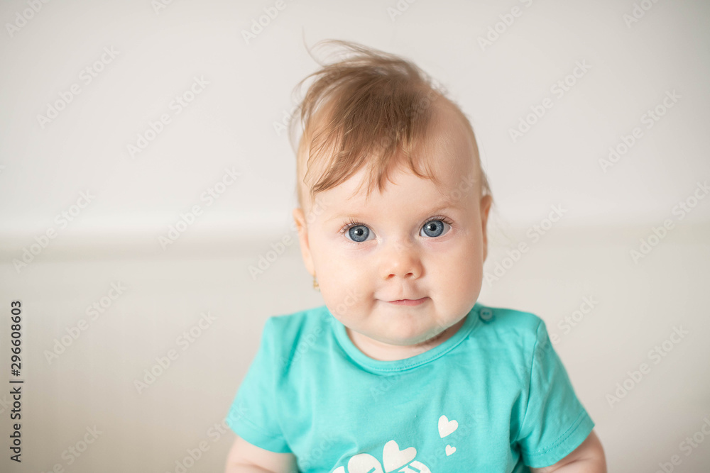 Portrait of adorable Caucasian baby girl with blue eyes, looking at the camera calmly, with curiosity, interactivity or inquisitiveness and sitting in a white baby cot; cute baby expressions concepts