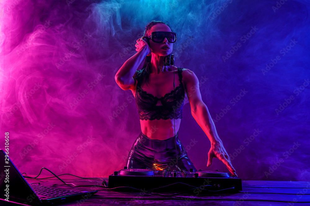 Young sexy woman dj in bra and sunglasses playing music. Headphones and dj mixer on table. Colorful Smoke on background - obrazy, fototapety, plakaty 