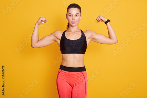 Close up portrait of young cute sporty brunette woman with ponitail, showing her arms and biceps to camera, female wears black bra and leggins. Sport, healthy lifestyle, women health, fitness concepts
