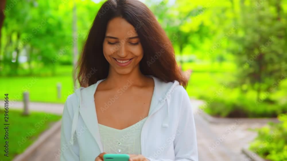 young woman wearing casual white hoodie walking chatting online or sharing post in social media outdoors background with green nature young woman wearing casual white hoodie walking chatting online or sharing post in social media outdoors background with green nature
