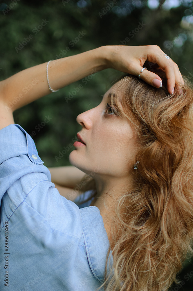 Naklejka premium Back view portrait of sexy blonde girl with curly hair wearing blue casual jeans blouse posing in the park on green trees background. Hipster style concept.