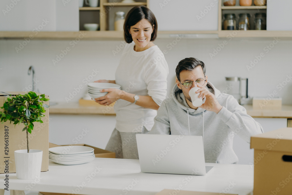 Busy husband works on laptop computer and drinks coffee, wears spectacles and casual sweatshirt, wife busy unpacking belongings, carries dishes, pose in kitchen during moving day. Family and mortgage Busy husband works on laptop computer and drinks coffee, wears spectacles and casual sweatshirt, wife busy unpacking belongings, carries dishes, pose in kitchen during moving day. Family and mortgage