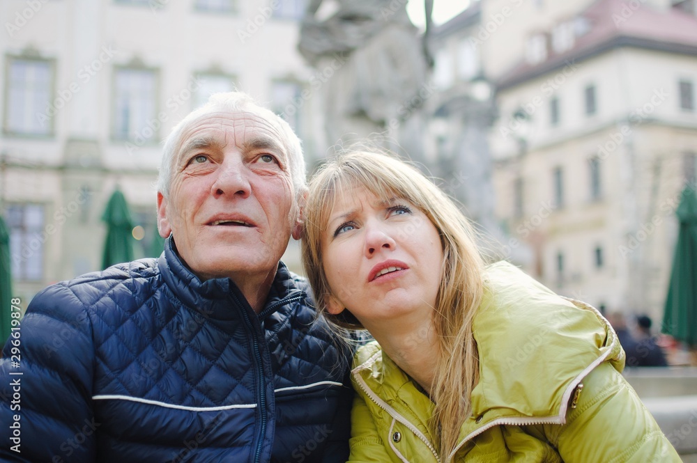 Outdoors portrait of elderly man and his young blonde wife spending time together in the ancient city during early spring or autumn. Couple with age difference pointing on some sightseeing place