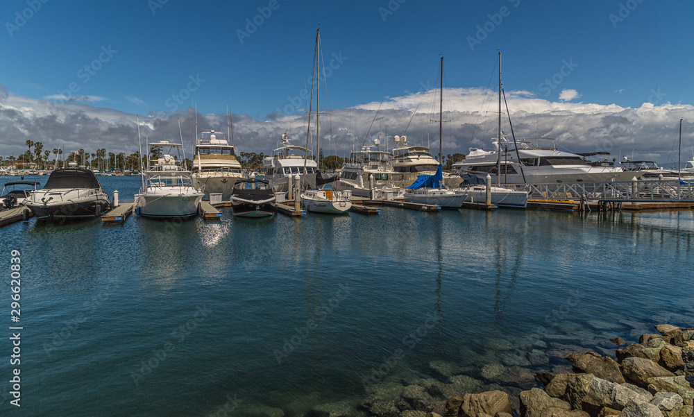 Yachts and boats lined up at the marina with rocks in foreground and clouds in sky in background