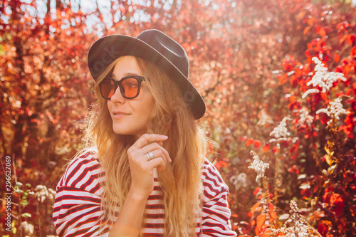 Portrait of a blonde in the autumn forest. Beautiful girl in a black hat walks in the park. Woman in stylish accessories outdoors. Portrait of a girl on the nature at sunset. red autumn background © Stanislav