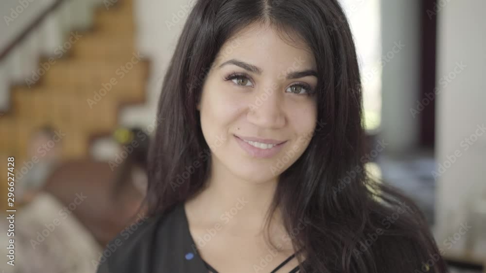 Close-up portrait of a happy beautiful Middle Eastern woman with hazel eyes and long dark hair looking at the camera and smiling. Her two little children playing at the background in cozy home. Close-up portrait of a happy beautiful Middle Eastern woman with hazel eyes and long dark hair looking at the camera and smiling. Her two little children playing at the background in cozy home.