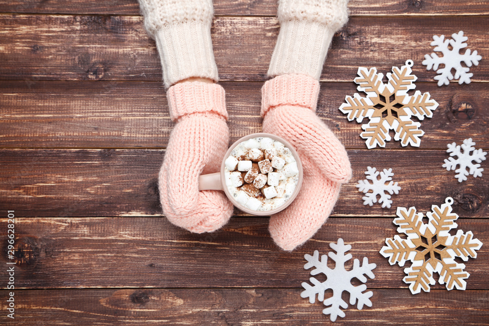 Hands in knitted mittens holding cup of hot drink with snowflakes on wooden table Hands in knitted mittens holding cup of hot drink with snowflakes on wooden table