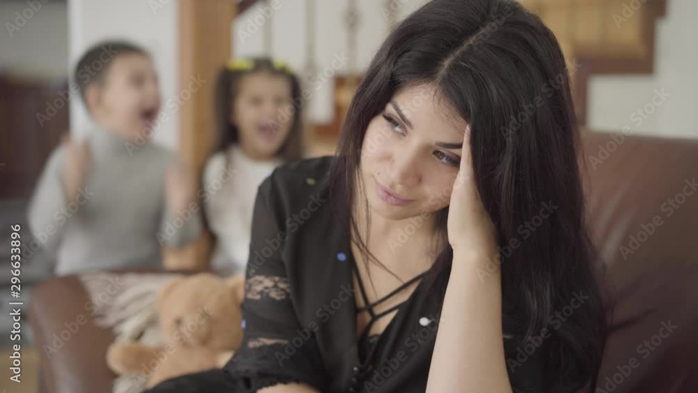 Portrait of a beautiful Middle Eastern mother with migraine sitting on the armchair and her two little children jumping at the background. Attractive woman holding her head tired from the noise. Portrait of a beautiful Middle Eastern mother with migraine sitting on the armchair and her two little children jumping at the background. Attractive woman holding her head tired from the noise.