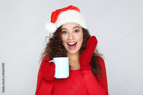 Beautiful american woman wearing santa hat and holding cup of tea on grey background