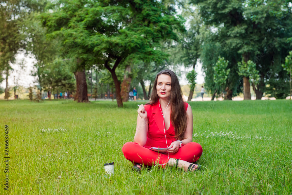 Portrait of young smiling business woman in red suit in earphones, doing online learning cources and writing in notebook while sitting on the green grass lawn in park. Self-development. Copy space. Portrait of young smiling business woman in red suit in earphones, doing online learning cources and writing in notebook while sitting on the green grass lawn in park. Self-development. Copy space.