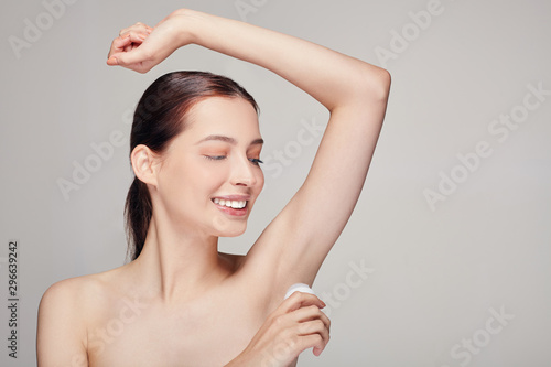 Happy pretty girl with brown hair with clean fresh skin posing on a gray studio background with deodorant in her hand near armpit looking side and smiling with wite teeth