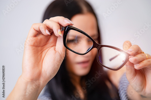Portrait of a young woman with spectacles in hands. Blurred white background. Girl looks through eyeglasses. Longhaired brunette beautiful girl and black framed eyeglasses. Closeup.