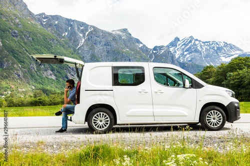 Young travel man sitting in car trunk talk on phone on nature mountains landscape on background.