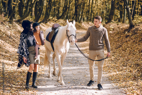 Young family is learning to ride a beautiful white horse in the autumn forest. Riding lesson