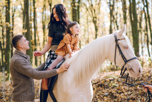Young family is learning to ride a beautiful white horse in the autumn forest. Riding lesson