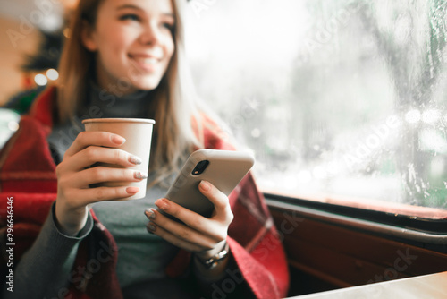 Smartphone and cup with coffee in the hands of a young happy girl spending time in a cafe. The girl writes a message on the phone and smiles. Focus on Smartphone. Background.