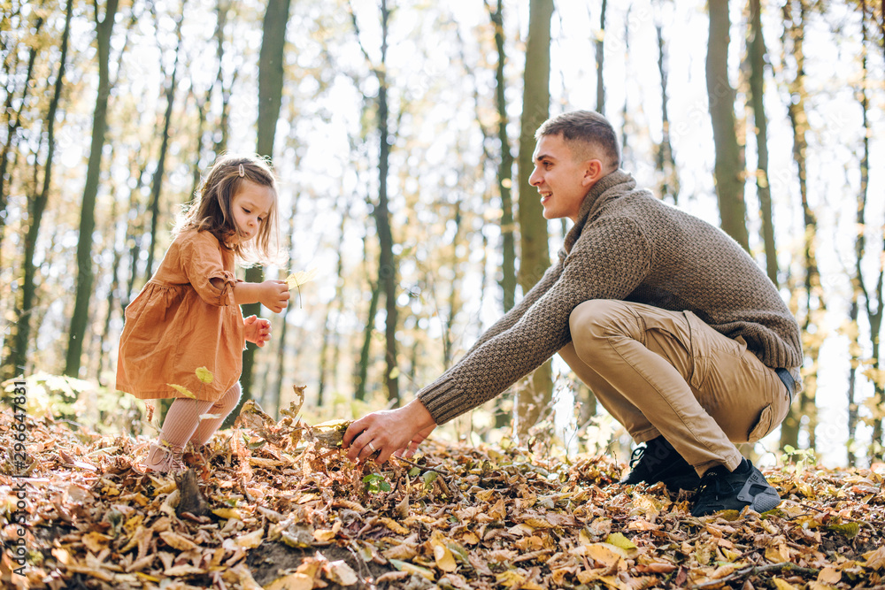 Fototapeta premium Family autumn weekend. Young father and his little daughter together in autumn park outdoors. Beautiful baby with leaves on top of dad.