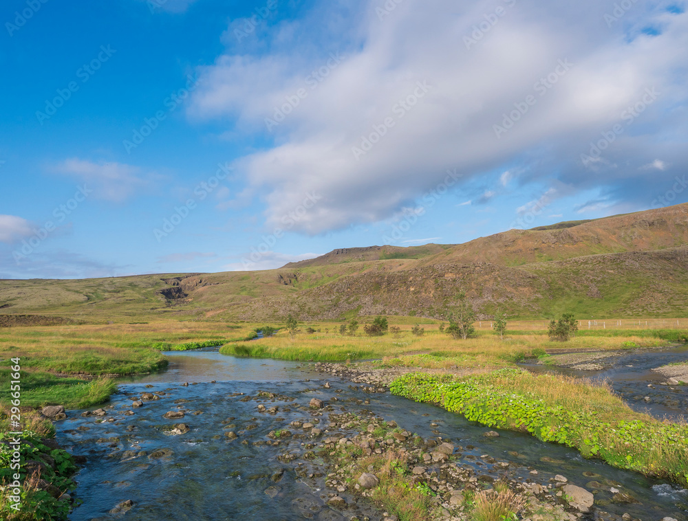Blue river stream, lush green grass meadow and hills at Reykjadalur hot springs area, South Iceland near Hveragerdi city. Summer sunny morning, blue sky.