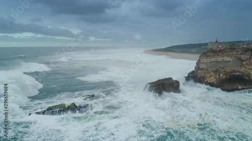 Aerial view of lighthouse on a cliff with a fortress on the coast of the Atlantic ocean with big waves at storm in Nazare, Portugal, 4k