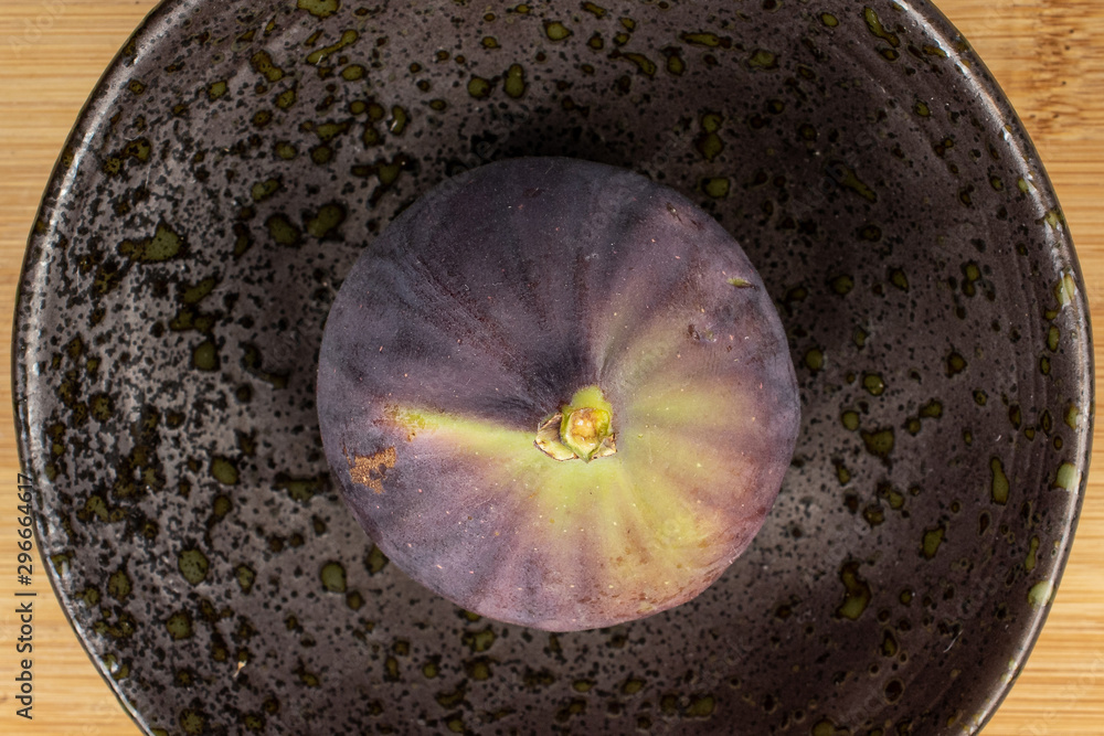 One whole sweet purple fig on glazed bowl flatlay on light wood