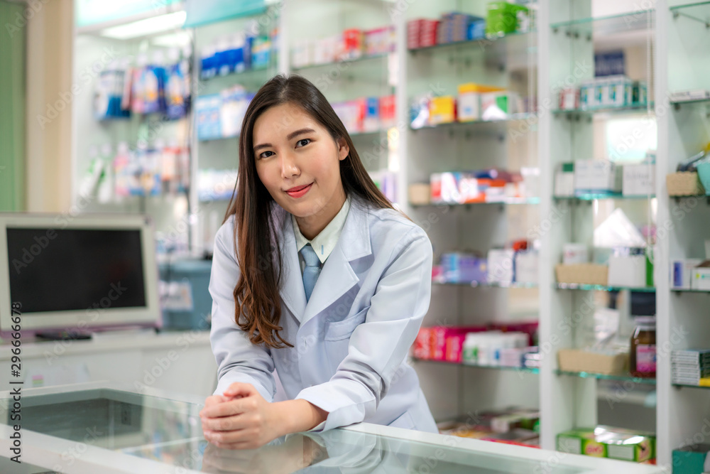 Confident Asian young female pharmacist with a lovely friendly smile standing leaning on a desk in the pharmacy drugstore. Medicine, pharmaceutics, health care and people concept.