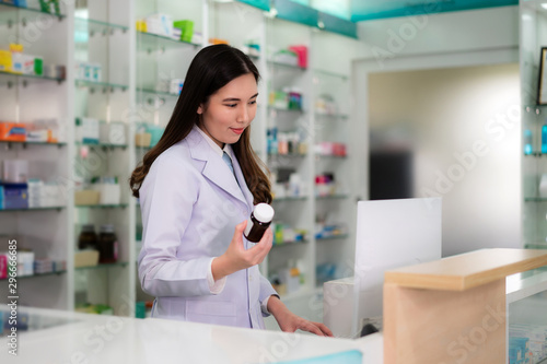 Confident Asian young female pharmacist with smile holding a medicine bottle and searching that product in the computer database in the pharmacy drugstore. Medicine, pharmaceutics, health care