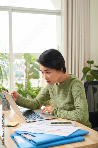 Young Asian woman sitting in office table, looking at laptop computer screen