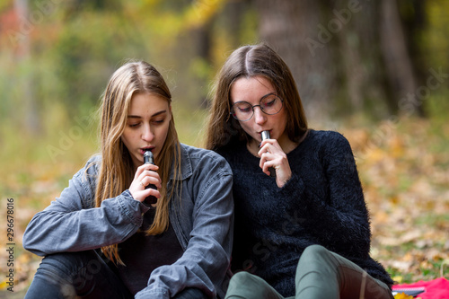 Vape lgbt teenagers. Bisexual lesbian young caucasian teenage girls in casual clothes vape electronic cigarette on the street in the park in the autumn evening. Bad habit.