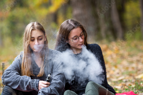 Vape lgbt teenagers. Bisexual lesbian young caucasian teenage girls in casual clothes vape electronic cigarette on the street in the park in the autumn evening. Bad habit.