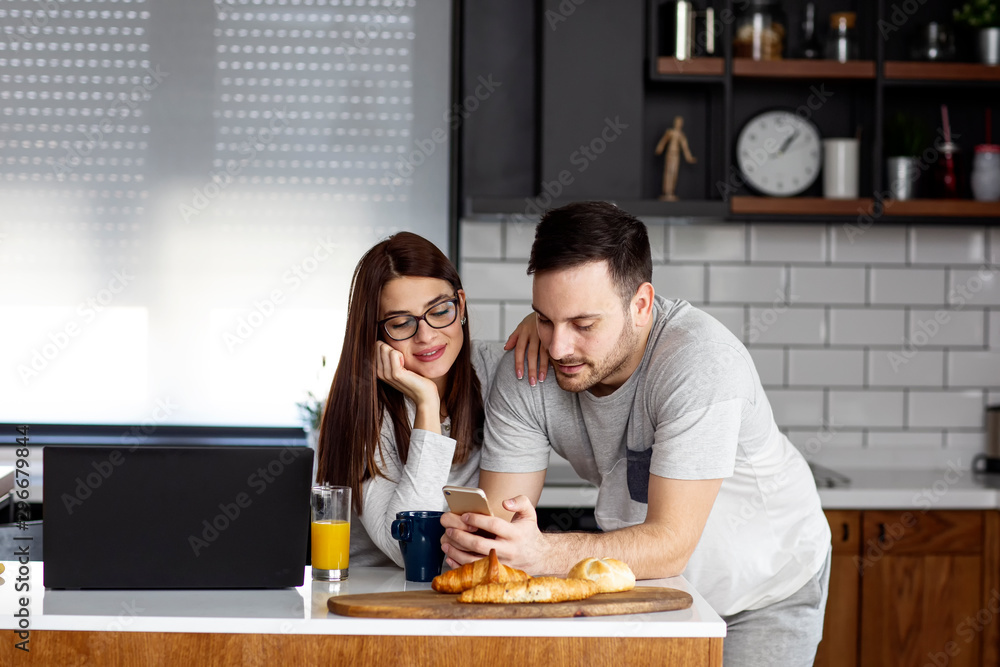 Fototapeta premium Young couple in pijamas eat breakfast at kitchen with laptop beside smile