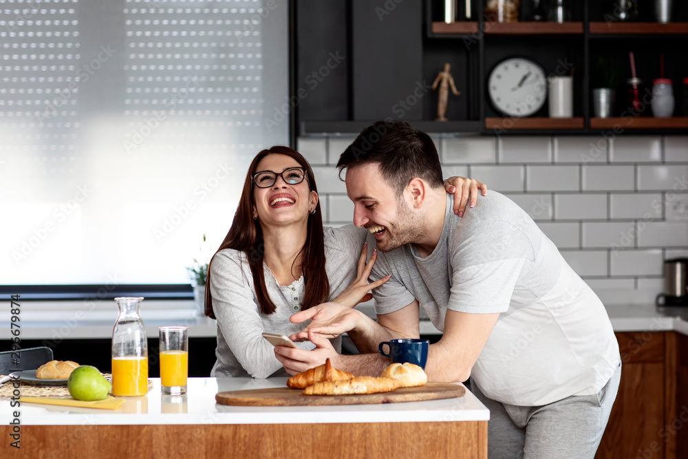 Fototapeta premium Couple in the kitchen eat breakfast with orange juice and pastry at table with mobile phone on hand smile