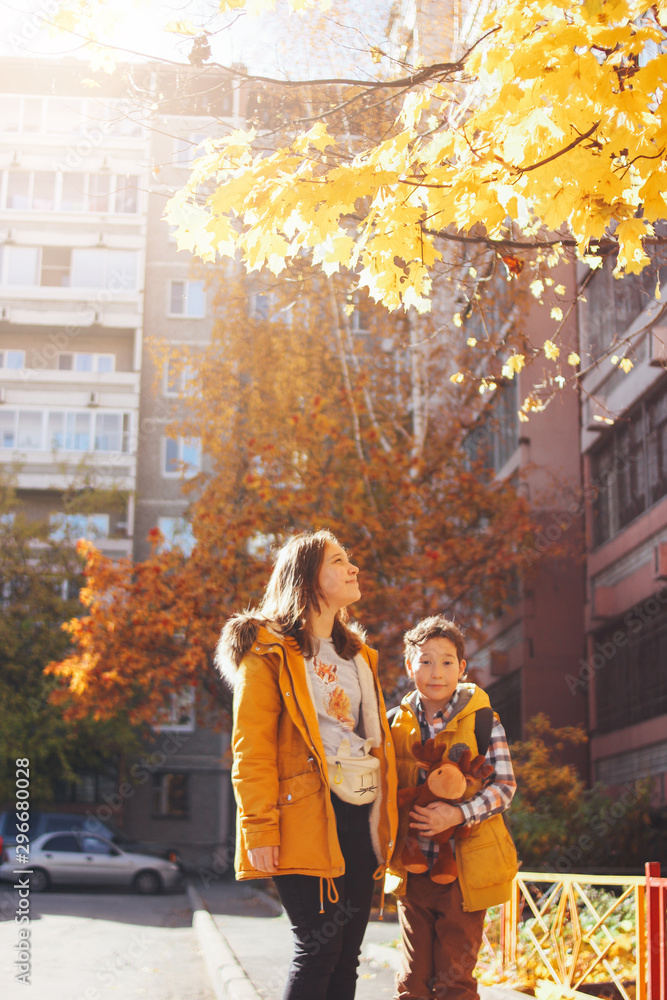 Fototapeta premium Smiling children brother and sister under sunny yellow tree maple in the courtyard of multi-storey building, golden autumn