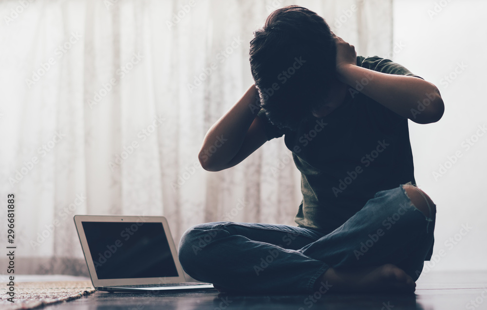 Cyberbullying concept. Young Asian preteen/teenage boy sit cross legged alone next to computer laptop in his room, covering his ears with hands. Stressed, frustrated, overwhelmed with online bullying. Cyberbullying concept. Young Asian preteen/teenage boy sit cross legged alone next to computer laptop in his room, covering his ears with hands. Stressed, frustrated, overwhelmed with online bullying.