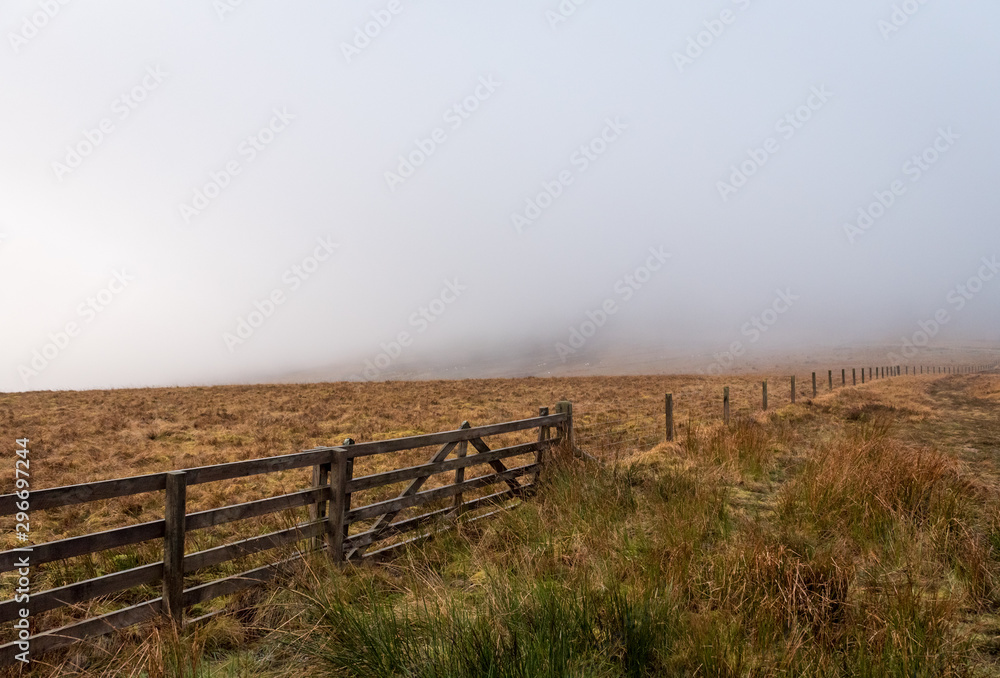 A wooden fence that stretches between Scotland and England border with brown grass field on either side on a foggy and grey day.