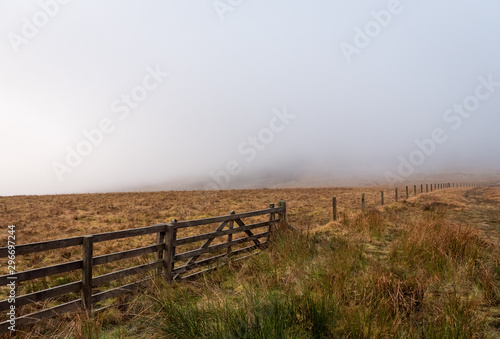 A wooden fence that stretches between Scotland and England border with brown grass field on either side on a foggy and grey day.