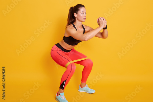 Fototapeta Naklejka Na Ścianę i Meble -  Studio shot of sporty woman squatting, doing sit ups with resistance band. Photo of Caucasian woman in fashionable sportswear isolated over yellow background. Strength and motivation concept.