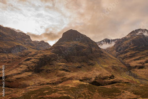 One of the three mountains called The Three Sisters  covered in brown wintery vegetation on a partially cloudy day in Scottish Highlands.