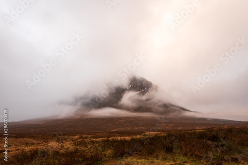 Wintery brown grass fields overlooks Buachaille Etive Mòr mountain covered in clouds in the Scottish Highlands.