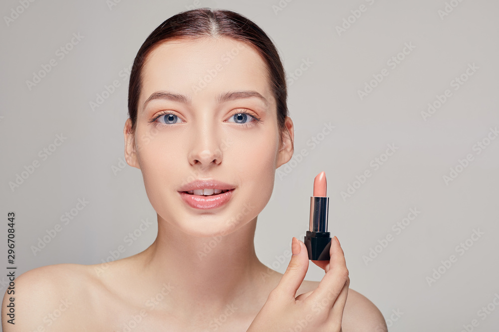 Beautiful girl with bare shoulders with lipstick in her right hand looks straight into the frame by her blue eyes with a slight smile and full lips Beautiful girl with bare shoulders with lipstick in her right hand looks straight into the frame by her blue eyes with a slight smile and full lips