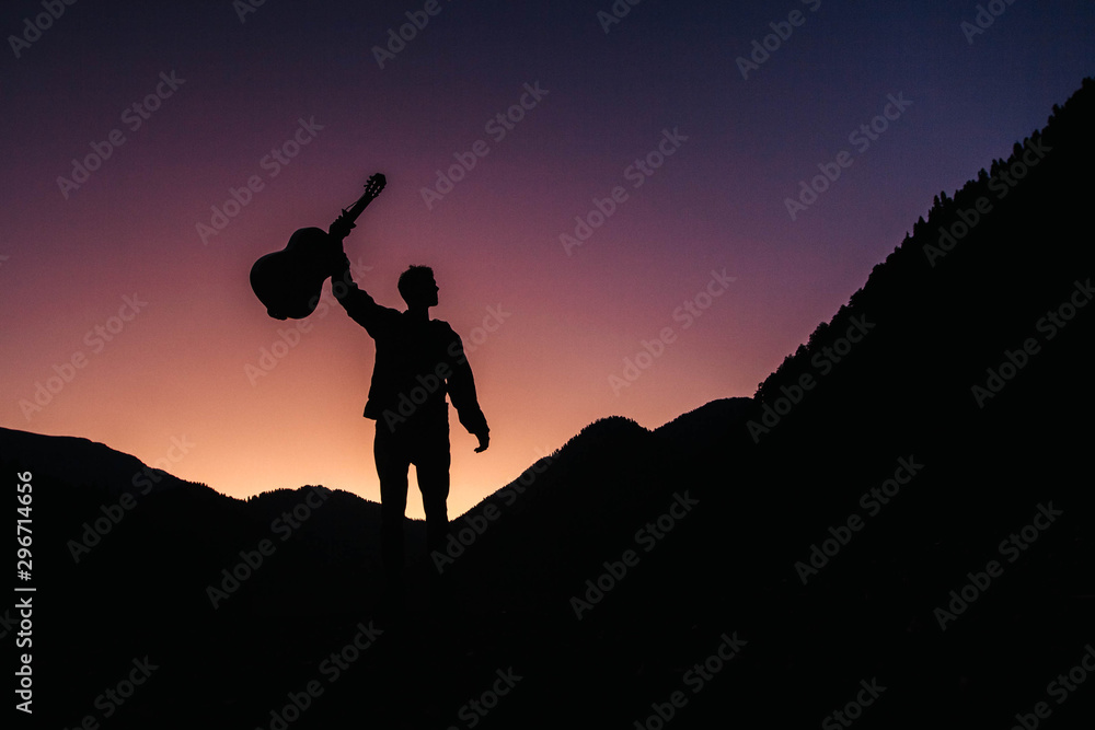 Silhouette of a man holding a guitar on a background of mountains and sunset. Concept of freedom relaxation. Place for text or advertising