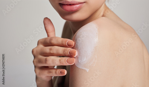 Close-up of a young girl with full lips and clean skin smears shoulder cream with cream on a light background. Dry skin care.
