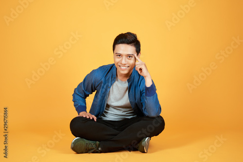 young casual asian man sitting with legs crossed and holding his head while smiling to the camera. isolated on color orange background © makistock