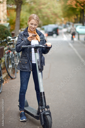 Young woman with electric kick scooter in the street of the city, looking at her phone and smiling. Public transportation renting service, modern urban ecological zero emission transport concept.